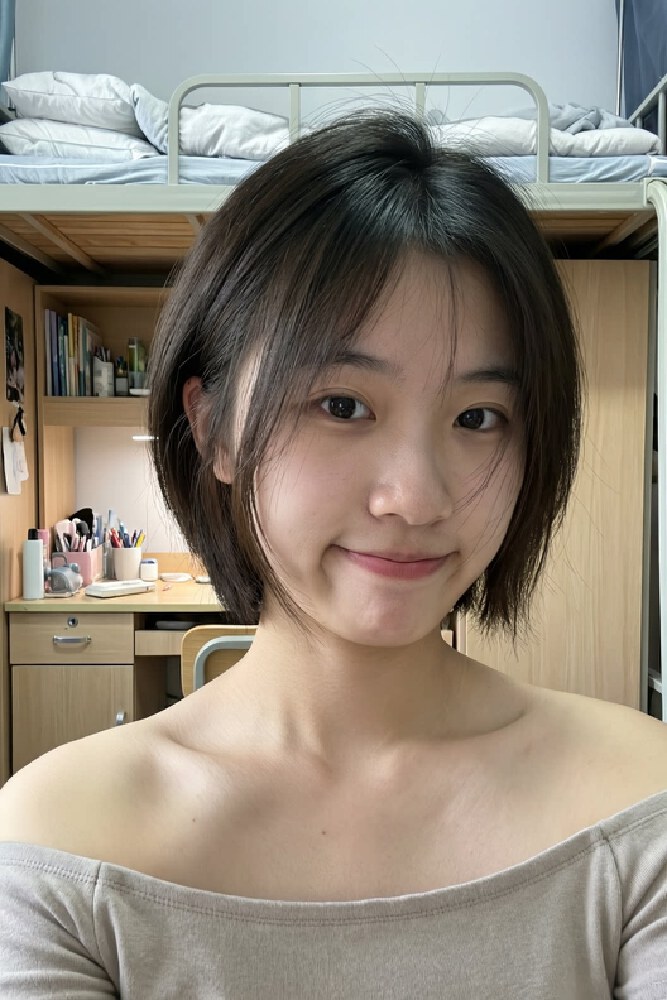 一位女生在宿舍内自拍，背景可见上下铺床、书桌及文具等物品