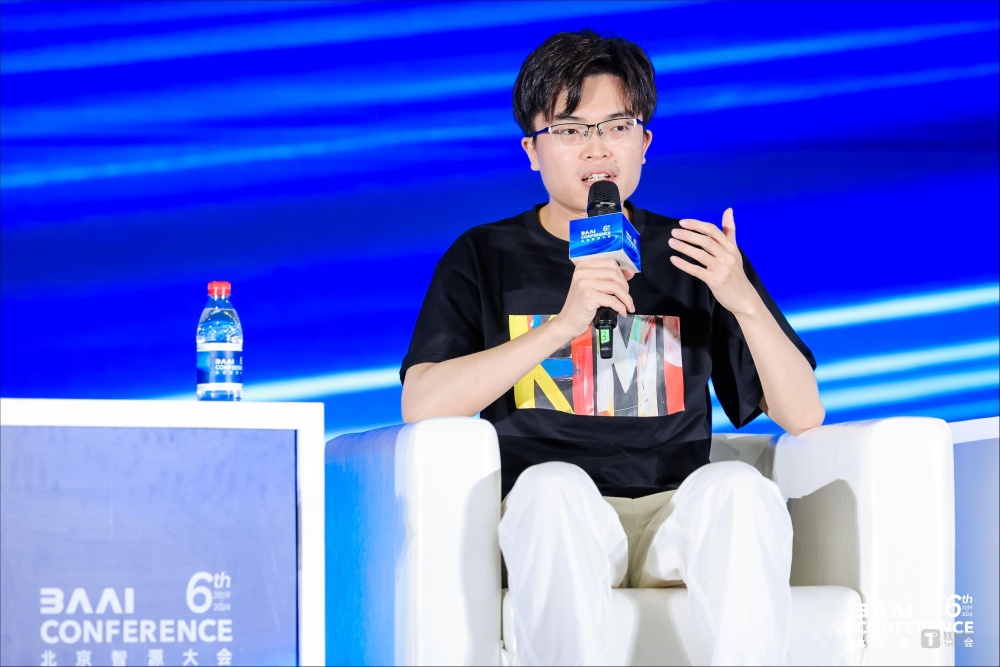 月之暗面大模型相关人物身着印有“KIMI”字样的黑色T恤，在“北京智源大会（BAAI CONFERENCE 6th）”活动中手持麦克风发言，背景为蓝色舞台幕布并带有会议标识，旁置矿泉水瓶。