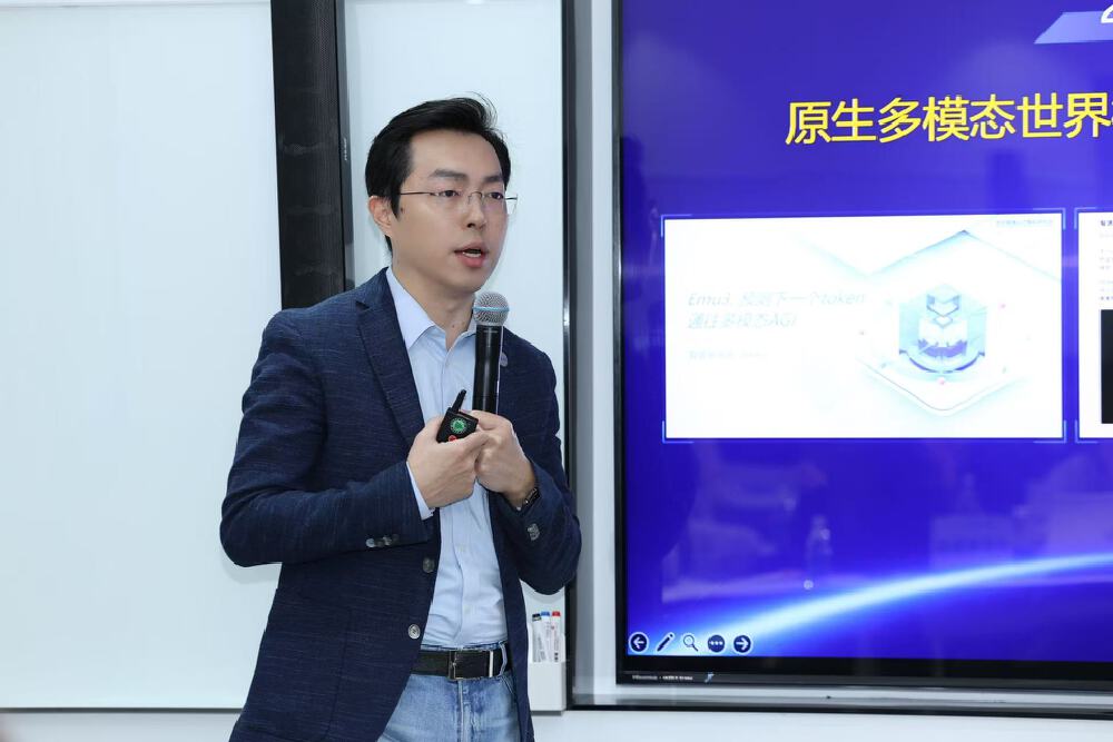 图片展示智源研究院发布会现场，一位人士手持麦克风演讲，背景屏幕呈现“原生多模态世界”及“Emu3 预测下一个token 通往多模态AGI”等多模态大模型技术相关内容，介绍其技术路线与产品。